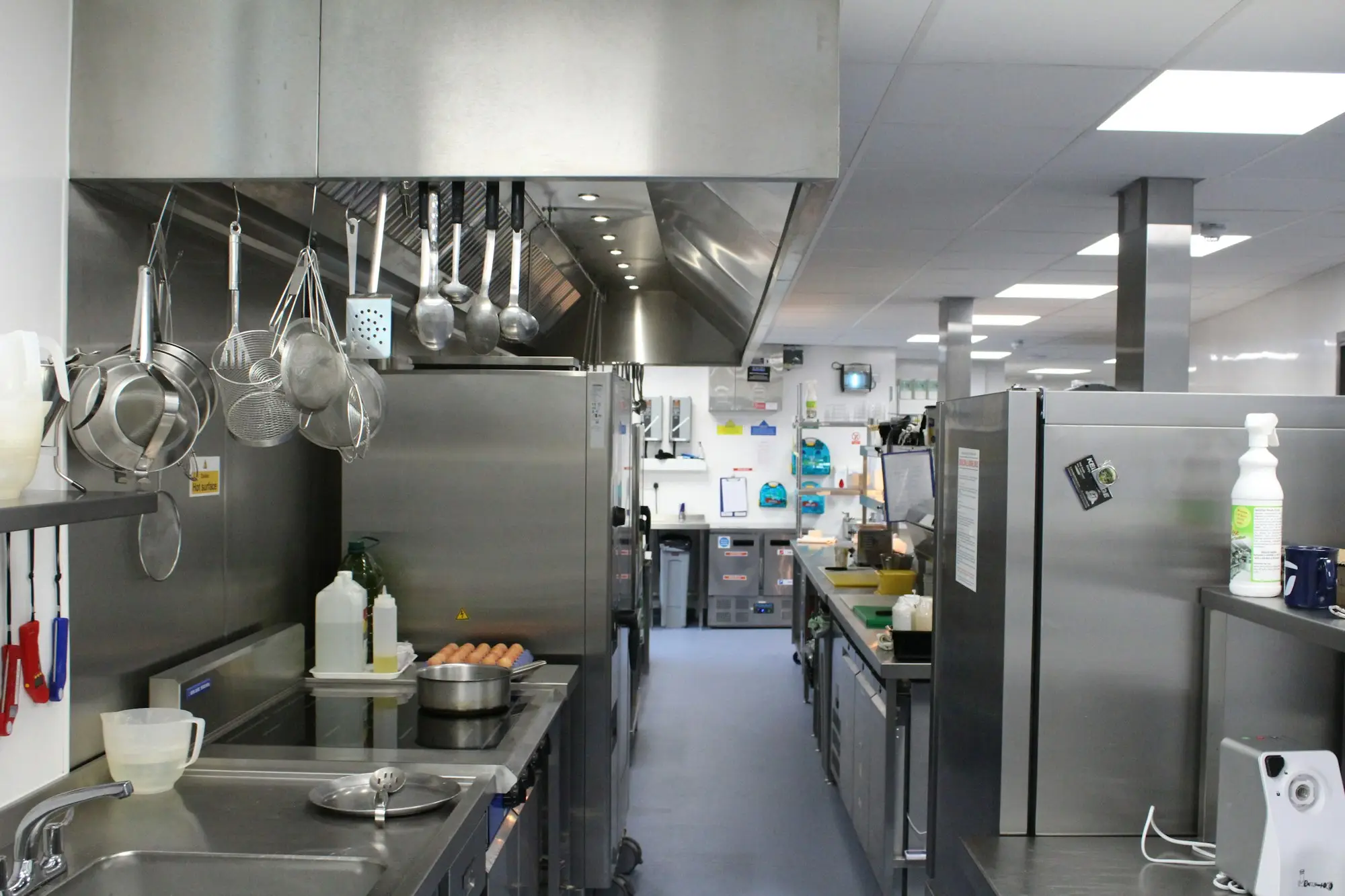 Stainless steel commercial kitchen with wash-up sinks in the foreground, hanging pans and an exhaust canopy over the cooking line