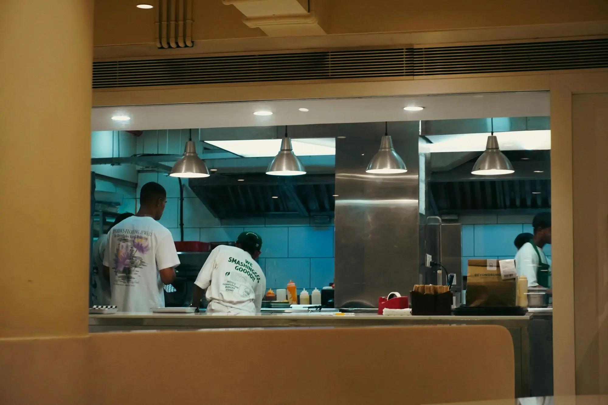 Cooks working under a stainless steel exhaust canopy in an open restaurant kitchen, seen across the pass