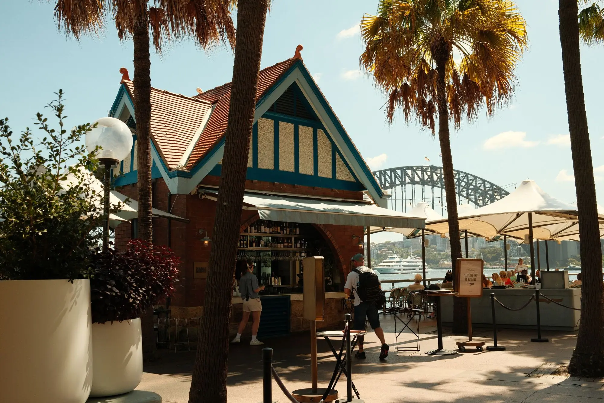 Waterfront café kiosk in Sydney with outdoor tables under white umbrellas, palm trees and the Harbour Bridge behind
