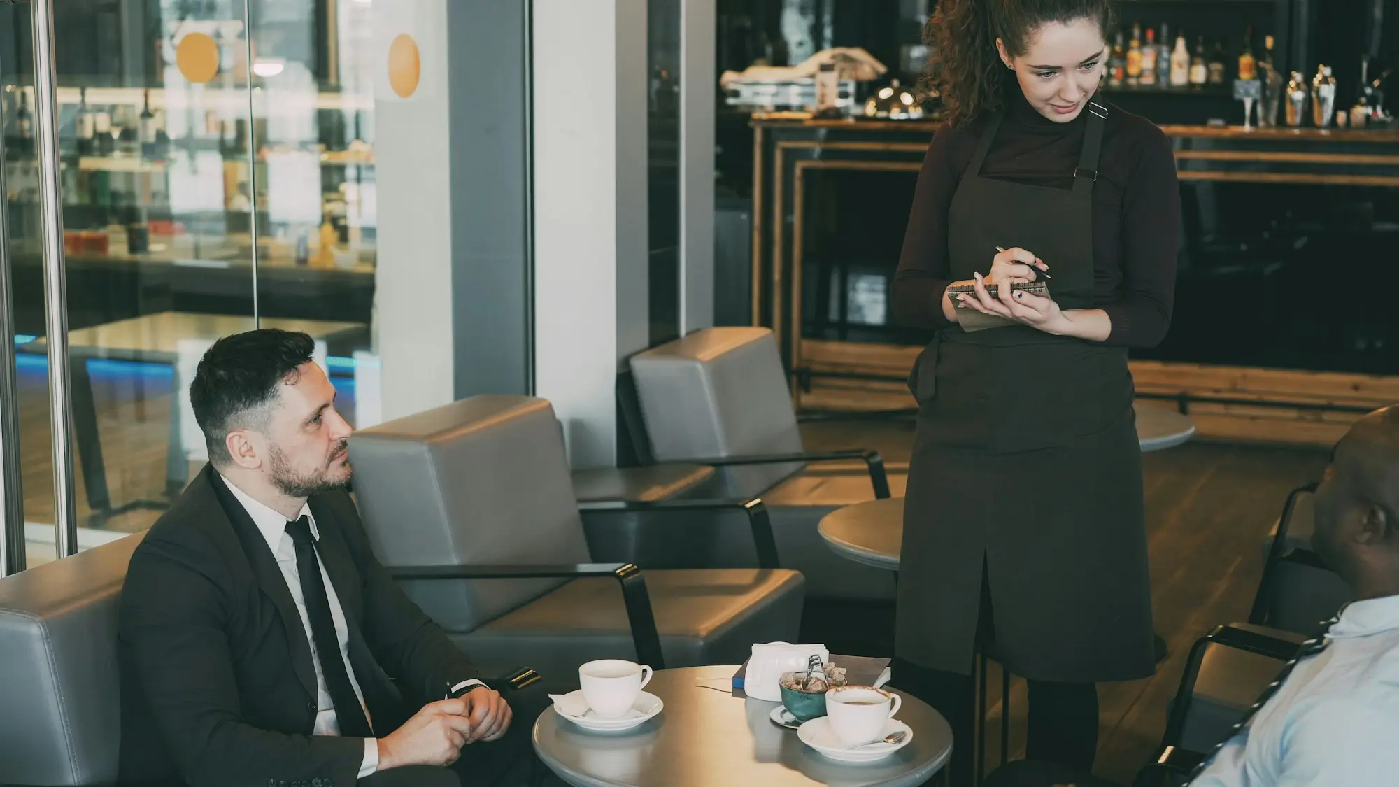 Waitress in a dark apron taking a customer's order at a café table, the moment a public holiday surcharge needs to be clear